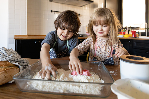 Little 3-year-olds playing with flour at home, helping mom prepare dough for bread. Happy children making animal figures on flour on the kitchen counter.