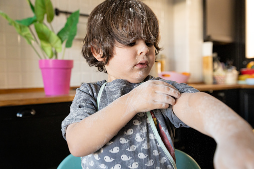 Dark-haired boy with a serious expression looking at his arm completely covered with flour. Little boy trying to clean flour from his clothes, in the background view of modern kitchen.