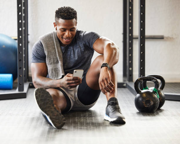 photo d’un jeune homme musclé utilisant un téléphone portable alors qu’il faisait de l’exercice dans un gymnase - technology mobile phone clothing smiling photos et images de collection