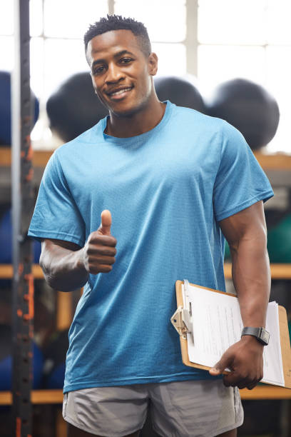 Portrait of a muscular young man showing thumbs up while holding a clipboard in a gym You won't go wrong by joining our gym gym men africa muscular build stock pictures, royalty-free photos & images