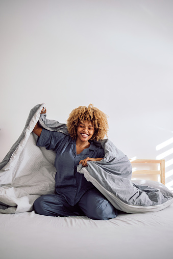 Woman in her pajamas smiling and sitting on the bed while playing with bed covers. She is holding them on her back like a cape.