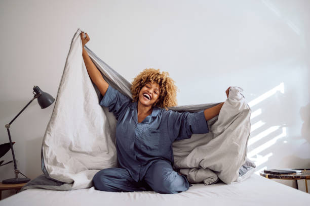 Happy Woman Playing with her Bed Covers in the Morning Woman in her pajamas smiling and sitting on the bed while playing with bed covers. She is holding them on her back like a cape. duvet stock pictures, royalty-free photos & images