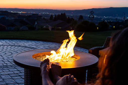 A young woman relaxes with her feet on a back yard fire pit enjoying the view from a hillside luxury back yard at sunset.