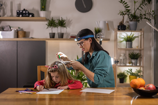 Mother wearing headlamp while cleaning lice from her little daughter's hair with comb, while she is drawing on paper at home.