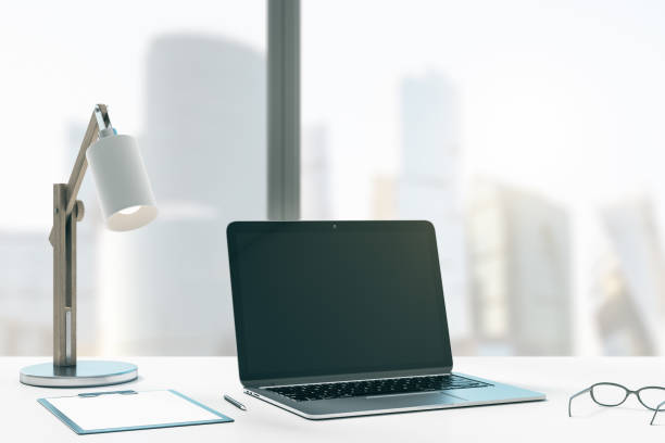istock Close up of creative designer office desktop with mock up place on black laptop computer screen, table lamp, supplies and other items. Blurry city view background. 3D Rendering. 1370786385