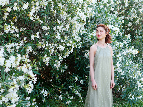 Portrait of young Asian woman standing in white oleander flower forest background, beautiful Chinese girl in green dress enjoy fresh air in forest park.