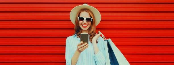 Happy smiling woman holding shopping bags and smartphone wearing summer straw hat and shirt on colorful red background stock photo