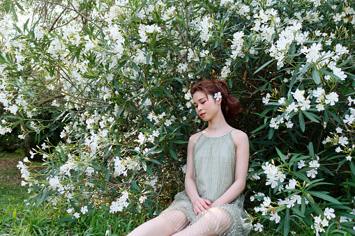 Portrait of young Asian woman sitting in white oleander flower forest background, beautiful Chinese girl in green dress enjoy fresh air in forest park.