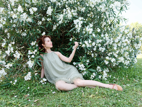 Portrait of young Asian woman sitting in white oleander flower forest background, beautiful Chinese girl in green dress enjoy fresh air in forest park.