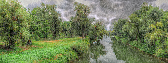 Natural landscape in the countryside with stream, trees and lush meadows during a summer thunderstorm with lightning on the horizon and raindrops