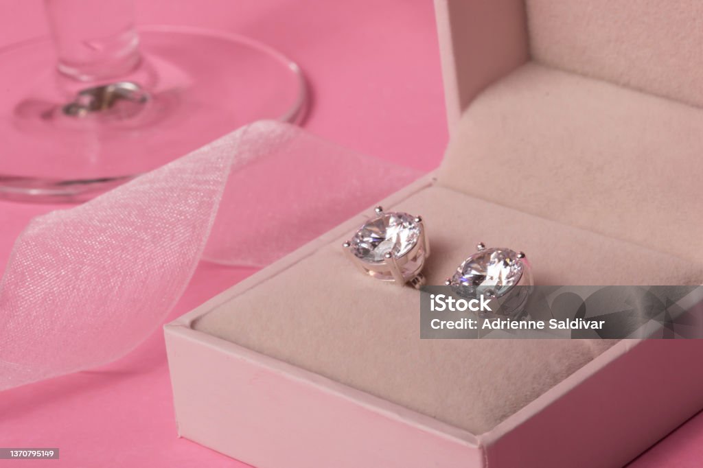 Diamond earrings in jewel box with champagne glass and ribbon on a pink background A pair of diamond stud earrings in a jewel box with a champagne glass and ribbon on a pink background. Romantic still life composition in pink monochrome color scheme. Diamond Earring Stock Photo