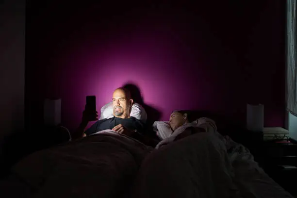 Man playing on mobile in bed while her wife sleeps next to him