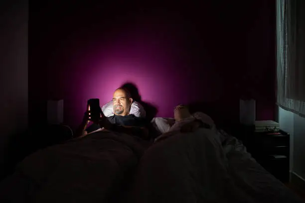 Man playing on mobile in bed while her wife sleeps next to him