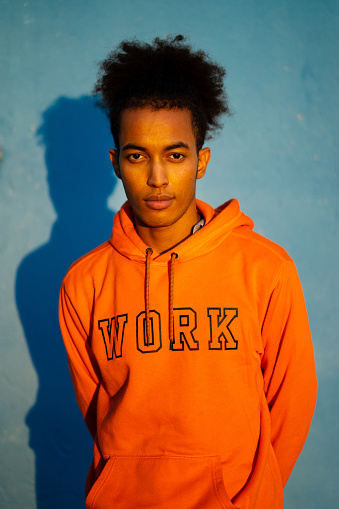 An African-American man with his hands behind his back standing against a blue background wearing an orange sweatshirt, looking seriously at the camera