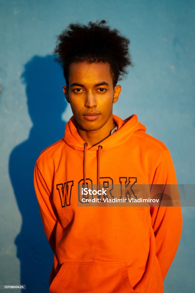 Man standing looking at camera An African-American man with his hands behind his back standing against a blue background wearing an orange sweatshirt, looking seriously at the camera Portrait Stock Photo
