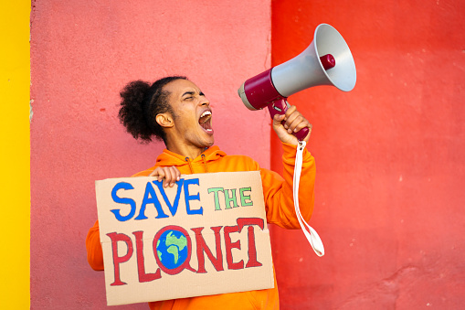 A Male conservationists holding a Save the Planet sign is using a megaphone to should his message loudly, standing against and stripped orange wall