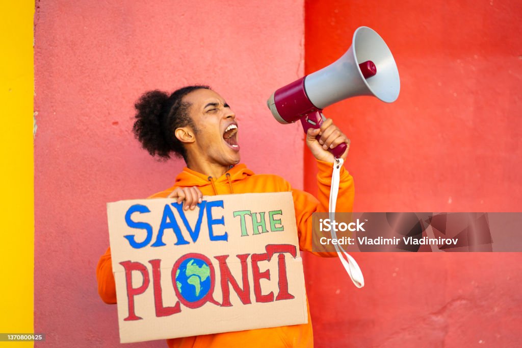 Protesting using megaphone against an orange striped wall A Male conservationists holding a Save the Planet sign is using a megaphone to should his message loudly, standing against and stripped orange wall Save The Planet Stock Photo