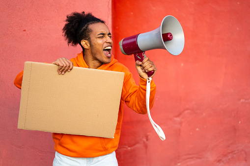 A multiracial activist holding a blank cardboard sign and shouting into a megaphone, he is wearing an orange sweatshirt and standing against orange walls