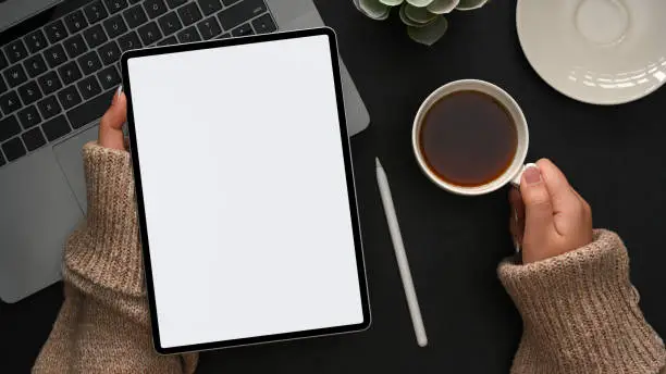 A female in comfortable sweater spending her free time on social media via tablet computer and sipping hot coffee. top view