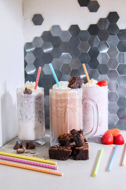 Glass mug of chocolate and strawberry and cookies and cream milkshake with rainbow straws on kitchen counter stock photo