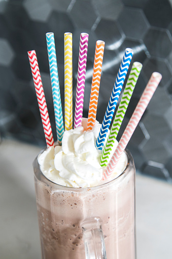 Glass mug of chocolate and strawberry and cookies and cream milkshake with rainbow straws on kitchen counter