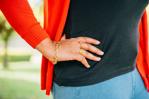 Fashionable women's accessories. Detail of the body of a model dressed in light blue jeans and a red jacket. Jewelry from golden ring and bracelet outdoor.