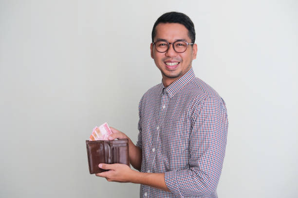 erwachsener asiatischer mann, der in die kamera lächelt, während er papiergeld aus seiner brieftasche nimmt - indonesian currency stock-fotos und bilder