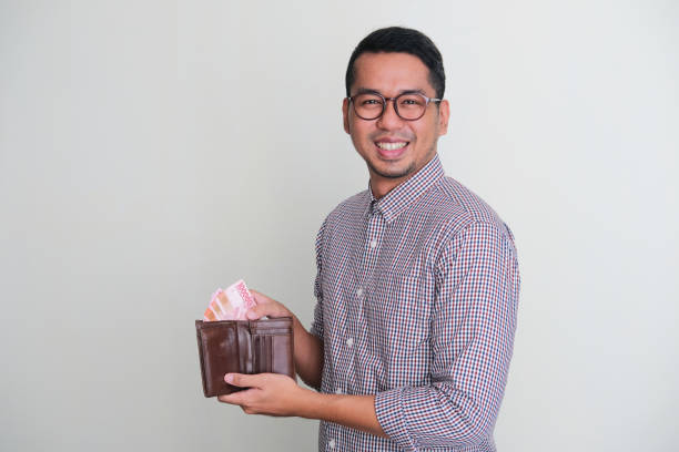 homme asiatique adulte souriant à la caméra tout en prenant du papier-monnaie de son portefeuille - indonesian currency photos et images de collection