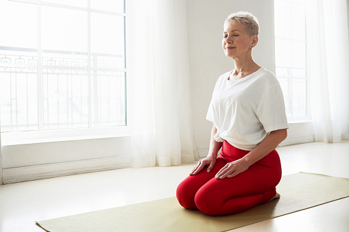 Spirituality, zen, zen and balance concept. Gray haired female on retirement sitting in virasana posture with eyes closed practicing meditation to reduce stress, imrove focus and concentration