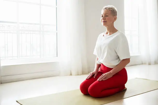 Photo of Spirituality, zen, zen and balance concept. Gray haired female on retirement sitting in virasana posture with eyes closed practicing meditation to reduce stress, imrove focus and concentration