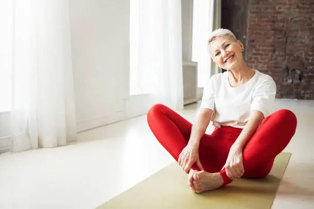 Photo of Sporty 50 year old gray haired woman sitting barefooted on yoga mat indoors doing bound ngle pose which helping to relieve symptoms of menopause. Aging, maturity, wellness and health concept