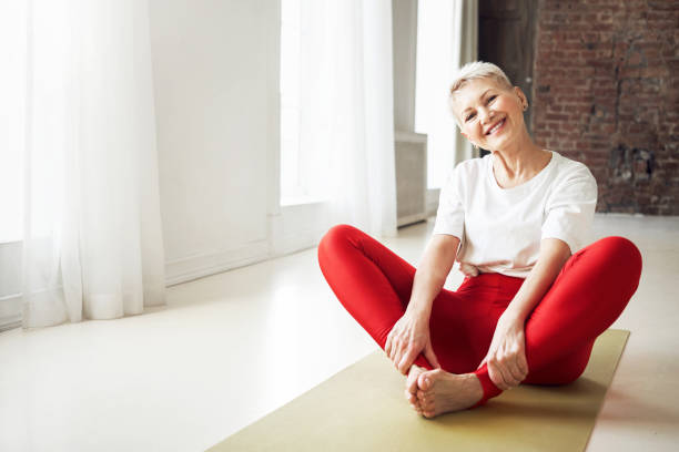 mulher de 50 anos de idade, de cabelos grisalhos, sentada descalça no tapete de ioga dentro de casa fazendo pose de ngle amarrada que ajuda a aliviar os sintomas da menopausa. envelhecimento, maturidade, bem-estar e conceito de saúde - healthy lifestyle yoga vitality flexibility - fotografias e filmes do acervo