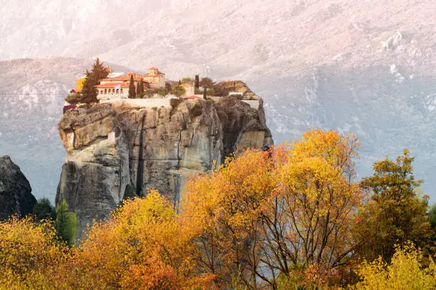 Atmospheric distant view of Monastery of Holy Trinity in Meteora with yellow autumn trees at foreground. Tourist and pilgrimage inspirations. Natural and religious wonders of Greece.