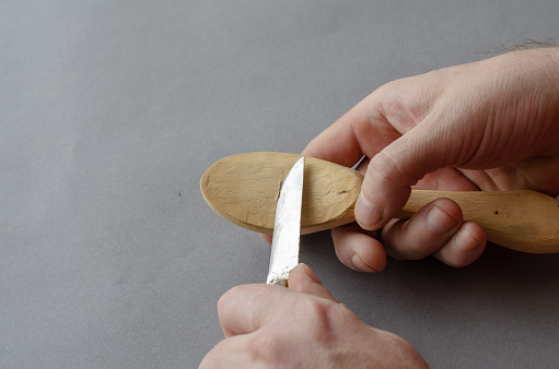 A man is planing a cormorant with a small knife against a gray background. The process of carving a wooden spoon. Selective focus.
