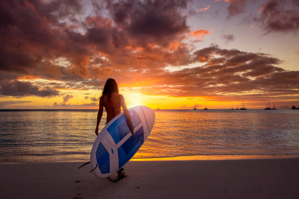 A sportive woman holds a Stand Up Paddle board on a tropical beach during a colorful sunset and is ready to enter the sea