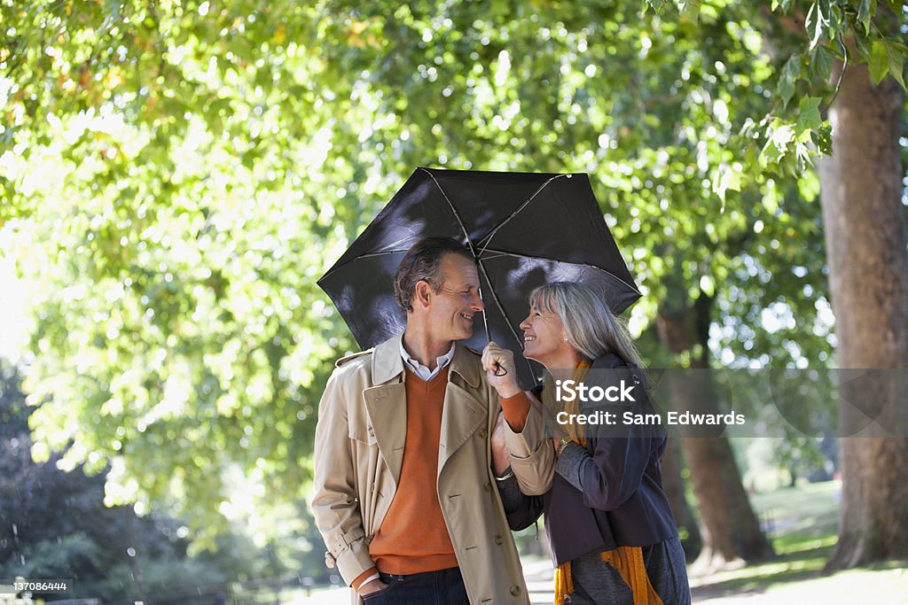 Coppia condividendo ombrello nel parco soleggiato - Foto stock royalty-free di 55-59 anni