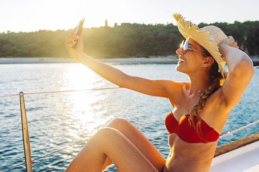 Young woman on yacht taking self portrait with mobile phone at sunset. She is very happy and attractive and she has a beautiful smile.