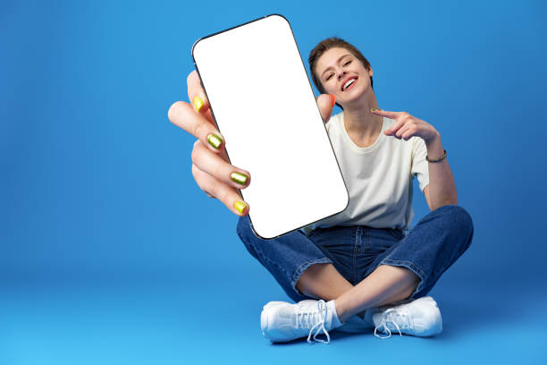 happy woman shows blank smartphone screen against blue background - mobiele telefoon stockfoto's en -beelden