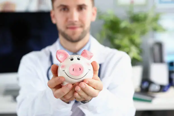 Portrait of man doctor hold white piggy bank, message to saveup money for healthcare. Doctor support savings for future, donation. Medicine, health concept