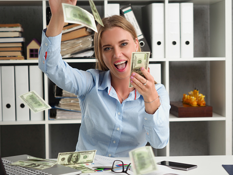 Portrait of cheerful woman throw in air dollars banknotes at work, cash everywhere in office. Business, big money, investment, profit, wealth, deal concept
