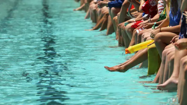 A bright aqua blue swimming pool with students sitting dangling their feet and toes in the water spectating. High school swimming carnival or club race meeting.