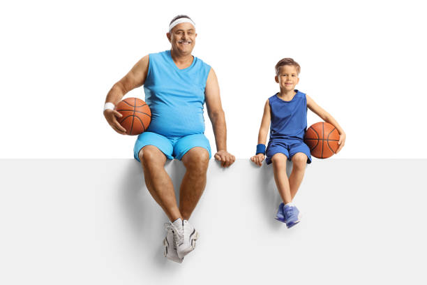 homme mûr en maillot tenant un ballon de basket et assis sur un panneau blanc avec un petit garçon - child basketball uniform sports uniform photos et images de collection