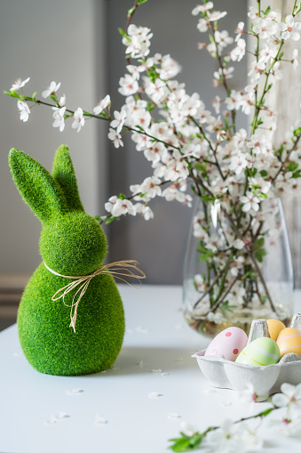 Green easter bunny with blooming flowers on tree branch in the vase and colored easter eggs in container on the white table. Happy Easter spring vertical card. Selective focus. Copy space