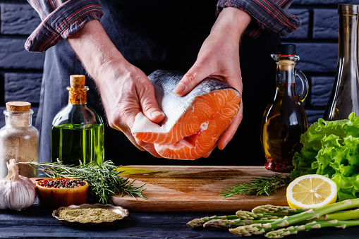 chef holding a piece of raw salmon fish on a restaurant kitchen. lettuce, rosemary, asparagus, lemon, bottles and spices on a with a black brick wall at the background