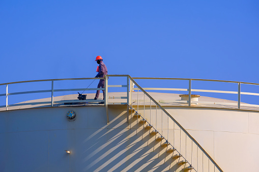 Engineer working to check oil quality on top of storage fuel tank against blue sky background