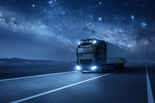 A commercial truck with a trailer is driving on a highway during nighttime. With a beautiful night sky above it.