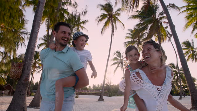 Happy parents and their small kids having fun while piggybacking on the beach in summer day.