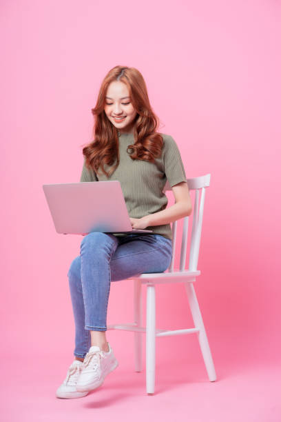 istock Young Asian woman sitting and using laptop on pink background 1370888082