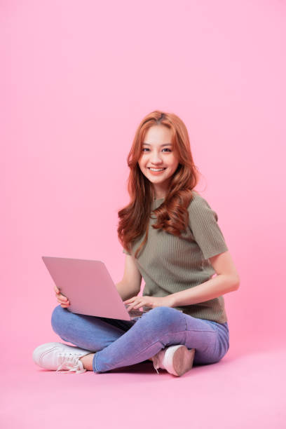 istock Young Asian woman sitting and using laptop on pink background 1370888178