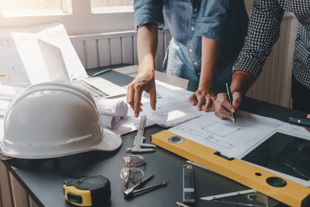 Close-up of architect and engineers holding a pen working on blueprints with safety equipment placed at the office. stock photo